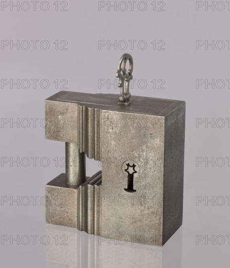 Padlock, 19th century. A padlock in the shape of a cuboid, the upper part topped with a kind of cornice; on each cornice two cuboids are connected by a cylindrical rod acting as an earring. After unlocking, the side wall together with the earring is pulled out, facilitated by an openwork handle on a hinge. The keyhole is horizontal on the longer wall. Materials/technique: locksmithing. Dimensions: height 12.6 cm, width 12.7 cm, depth 6.2 cm., A metal rectangular block with a matte silver-gray surface stands upright against a pale gray background, showing worn and slightly mottled textures and a few darker spots; the block has a vertical slot creating an open gap in its left side revealing a cylindrical metal rod with a similar silver-gray finish, and the edges of the slot are fluted with a ridged pattern; on top of the block a decorative metal loop with an ornate shape and a darker pewter tone is attached, and the block casts a faint reflection on the glossy surface beneath it.