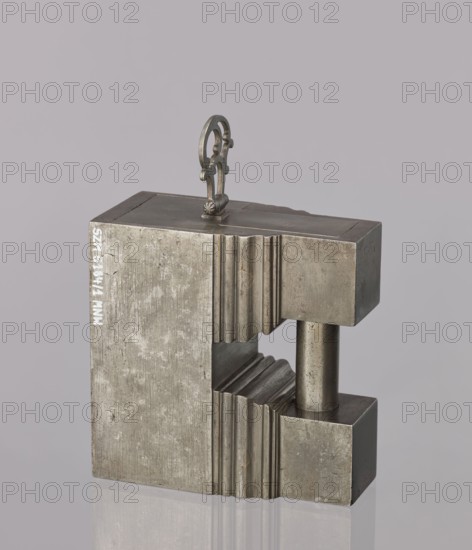 Padlock, 19th century. A padlock in the shape of a cuboid, the upper part topped with a kind of cornice; on each corner two cuboids are connected by a cylindrical rod acting as an earring. After unlocking, the side wall together with the earring is pulled out, facilitated by an openwork hinged handle. The key hole is horizontal on the longer wall. Maker, locksmithing. Height 12.6 cm, width 12.7 cm, depth 6.2 cm., A metallic block-like object with a matte silvery-gray surface stands against a pale gray background. The object has a rectangular, geometric shape with a cutout section revealing a cylindrical metallic rod connecting the top and bottom parts. Both the inner and outer faces show vertical ridged grooves and smooth flat surfaces with a slightly worn, textured finish. A small ornamental metal handle with a looped decorative design is attached to the top surface. The overall colors visible are silvery-gray and pale gray.