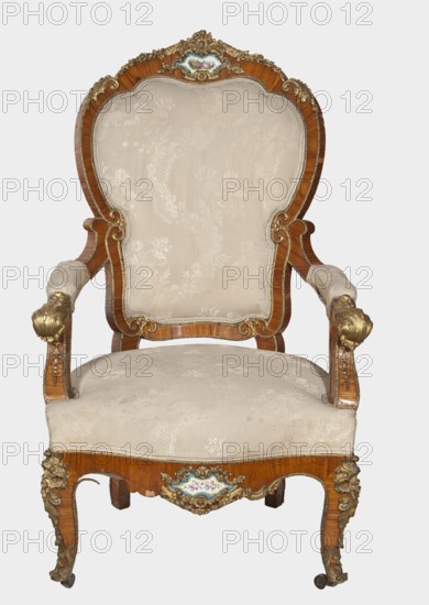 Armchair, origin unknown, neo-Rococo rocaille ornament, decorated with flowers, acanthus ornament, fêtes galantes, campanulas, and floral bouquets. Upholstered backrest, seat, armrests, veneered with rosewood, decorated with porcelain plaques and cast brass fittings. S-shaped legs widening in the upper section cabriole, arching into a frame with a wavy, bulging outline; front ends with rollers. Armrests repeat the frame outline, on recessed slightly S-shaped supports, topped with volutes. Backrest in the form of a symmetrical cartouche widening upwards, framed by channel sections topped with volutes; with a clearance. Porcelain plaques painted with flowers set in the center of the front frame and backrest finial, framed by brass composed of rocaille, flowers, and acanthus leaves. Frame edges accented with brass strips; brass fittings include a garland at the backrest finial, volutes at armrest ends, acanthus plaques and slippers at knees and feet. Seat upholstery on springs; upholstery...