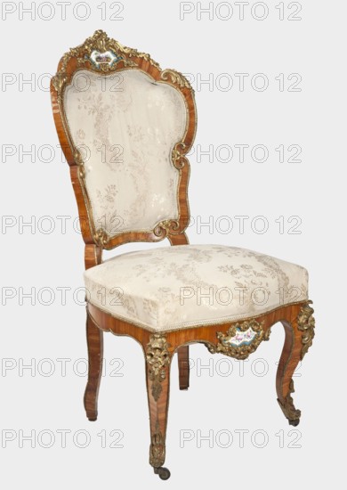 Chair, origin unknown, neo-Rococo Neo-Rococo CHAIR, rocaille ornament, flowers, acanthus ornament, fêtes galantes, floral bouquets, cartouches. Chair with padded backrest and seat, veneered with rosewood, decorated with porcelain plaques and cast brass fittings. S-shaped legs, widening in upper section, arching into a wavy, bulging frame; front ends with rollers. Backrest in form of a symmetrical cartouche, widening upwards, framed by channel sections topped with volutes; with a clearance. Porcelain plaques painted with flowers set in brass frames composed of rocaille, flowers, and acanthus leaves at center of front frame and backrest top. Frame edges accentuated with brass strips; knees and feet feature acanthus plaques and slippers. Seat upholstery on springs; upholstery fabric not original, with lace ribbons featuring floral bouquets. Date: 1850–1870, likely France. Seating furniture; height 103 cm, width 54.5 cm, depth 66.5 cm., A single ornate wooden chair with a curved high back ...