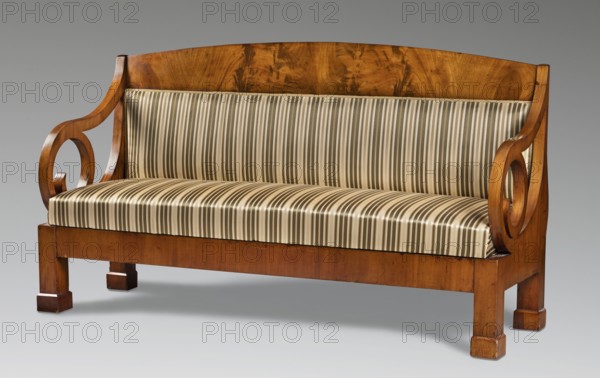Sofa, Biedermeier style, ca. 1820–1830. Upholstered seating piece seat, backrest with armrests that, together with the supports, form volutes of considerable diameter; supported by four angular straight legs. Rectangular flat frame; rectangular seat with applied spring upholstery on the frame. Flat backrest topped with a board with a slightly arched top. Side stiles and armrests have square cross-sections. Between them and the lower part of the volutes are arched boards forming spherical triangles. Reupholstered in green striped fabric. Height 104.5 cm; width 186 cm; depth 62.9 cm. Place of origin possibly Warsaw., A wooden settee with a warm brown polished finish and a gently curved backrest, featuring decorative open scroll armrests on each side; the seat and back cushions are upholstered in striped fabric showing shades of beige, cream, light brown, and pale gray; the settee stands on square block legs in the same brown wood, and the image background is a smooth neutral gray.