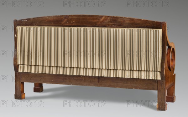 Sofa, Biedermeier style, upholstered sofa seat, backrest with armrests that, together with the supports, form volutes of considerable diameter, supported by four angular straight legs. Rectangular, flat frame; rectangular seat with applied spring upholstery on the frame. Flat backrest topped with a board with a slightly arched top. Side stiles and armrests have a square cross-section. Between them and the lower part of the volutes are arched boards forming spherical triangles. Reupholstered with a green striped fabric. Dated 1820–1830, possibly Warsaw. Piece of furniture seating piece. Dimensions: height 104.5 cm, width 186 cm, depth 62.9 cm., A wooden bench with a rectangular back panel upholstered in vertically pleated fabric, showing a group of beige and cream tones with narrow darker beige stripes; the frame is medium to dark brown wood with visible grain and some scuffs and lighter worn areas, including curved arm supports on the sides and squared legs; the background is a smooth ...