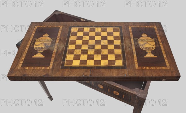 Game table, games, classicism style, rosette ornament, chessboards, vases, classicist. Rectangular game table, square when unfolded, supported by four angular, converging legs with wedge feet; veneered and inlaid. The top is divided into three rectangular fields, enclosed by a ribbon and two veins, filled with vases outermost and a chessboard. 18th/19th century, Central Europe, possibly Gdansk. Furniture; game furniture. Height 77 cm, width 42.5 cm, depth 86 cm., A wooden tabletop with a decorative inlaid chessboard at its center and a decorative panel on each side, all in tones of brown, tan, beige, and yellow, set against a pale gray background; the central chessboard shows alternating light and dark wood squares, the side panels each feature an inlaid urn-like motif in lighter wood against a darker brown field framed by a lighter border, with visible wood grain, varnish sheen, and some worn or scuffed areas; beneath the top, parts of a wooden frame and legs are visible in dark brown...