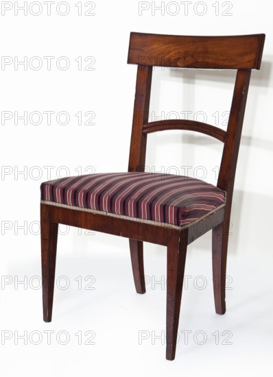 Chair, furnishings of representative buildings of the Republic of Poland 1918–1939, Biedermeier, Classicism, mahogany chair with a padded seat and a board-and-openwork backrest, supported by four angular, converging legs — front straight, rear arched outwards. Trapezoidal seat, straight frame. Backrest angled outwards, with stiles extending the rear legs, fastened with an arched, profiled strip and a crowning board curved in line with the back. Upholstery connected to the frame. 1815–1830, Warsaw; seating furniture. Height 87.3 cm, width 46.5 cm, depth 43.5 cm., A wooden chair with a dark brown finish and visible wood grain stands against a light background; it has a straight rectangular top rail and a simple curved slat below it connecting the back uprights, tapered legs, and an upholstered seat covered in a striped fabric with shades of burgundy, pink, black, and gray, edged with a narrow decorative trim.