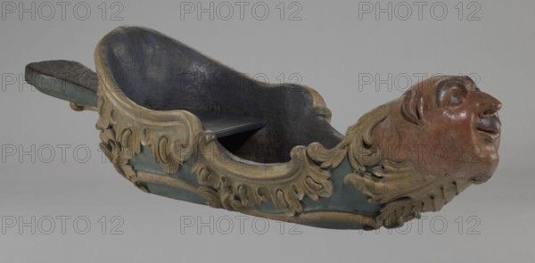 Sleigh, rococo two-seater, carved, polychrome, gilded; 3rd quarter of the 18th century, Poland . The sleigh is shaped like a gondola made using the dugout canoe method, painted blue, with a wavy upper rim highlighted by a half-cylinder and channel sections with rocaille motifs. The front is a mascaron painted red, framed with acanthus leaves; at the rear is a trestle, originally upholstered, with carved acanthus beneath. Inside the sled are four sockets for connection with skid supports. Height 57 cm, width 208 cm, depth 68 cm., A sculpted wooden object shaped like a shallow boat with a carved humanlike head at one end and an extended flat tail at the other, featuring ornate scroll and leaf carvings along the sides. The interior of the boat is dark, appearing black or very dark blue, while the exterior surfaces show muted teal and faded gold tones on the decorative elements and trim. The carved head has reddish brown and pinkish brown hues with darker brown shading around facial featur...