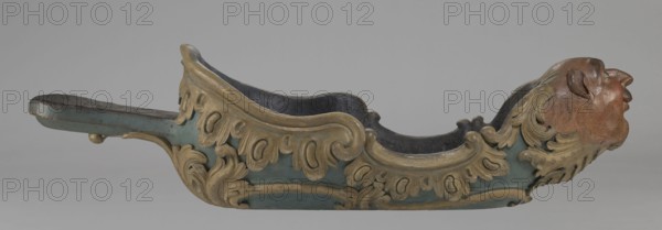 Sleigh, rococo two-seater, carved, polychrome, gilded, 3rd quarter of the 18th century, Poland . The sleigh is in the shape of a gondola made using the dugout canoe method, painted blue, with a wavy upper rim highlighted by a half-cylinder and channel sections with rocaille motifs. The front is in the form of a mascaron painted red, framed with acanthus leaves. At the rear is a trestle, originally upholstered, with a carved acanthus underneath. Inside the sleigh are four sockets for connection with skid supports. Vehicle; height 57 cm, width 208 cm, depth 68 cm., A carved wooden object shaped like a long curved trough with ornate scrollwork and a sculpted humanlike head at one end; the body features raised decorative leaf and curl motifs in a goldish color over a muted teal base, with the interior appearing dark, and the head showing natural wood tones with reddish-brown and tan hues and carved facial features and hair.