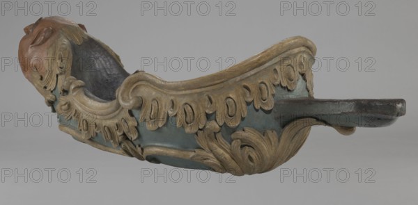 Sleigh, rococo two-seater, carved, polychrome, gilded; 3rd quarter of the 18th century, Poland . The sleigh is in the shape of a gondola made using the dugout canoe method, painted blue, with a wavy upper rim highlighted by a half-cylinder and channel sections with rocaille motifs. Front in the form of a mascaron painted red, framed with acanthus leaves; rear trestle originally upholstered with carved acanthus underneath. Inside the sled are four sockets for connection with skid supports. Subject elements: mascarons, men's heads; ornaments: rococo, rocaille, acanthus. Dimensions: height 57 cm, width 208 cm, depth 68 cm., A carved ornamental object with ornate curving leaflike motifs and scrollwork, showing a sculpted head at one end and a shallow flat extension at the other, painted in muted blue, beige, brown, and hints of dark gray and black, all set against a plain light gray background.