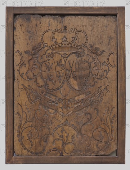 Leather embossing mold, creator unknown, rococo style, Wettin family panoply ornament, Order of the White Eagle, Polish-Lithuanian Commonwealth coat of arms, dated 1730–1760, possibly Poland or Saxony; height 96.5 cm, width 73 cm, depth 4.2 cm., A rectangular wooden panel with a darker wooden frame around it, showing an ornate carved design; the main surface is medium brown with darker brown carved lines forming a central heraldic motif topped by a carved crown and flanked by two winged cherub-like figures, with swirling floral and foliate scrollwork filling the lower area. The carving includes shapes resembling shields, crossed objects, draped banners, and stylized flowers and leaves. The wood shows variations in tone with lighter brown areas, darker brown spots, and signs of wear and small abrasions along the top edge and corners. The background behind the framed panel is a pale gray.