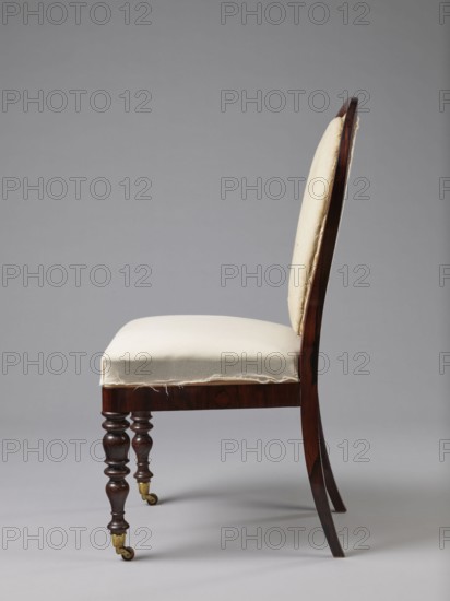Chair, ca. 1840, Warsaw. Seating furniture with a padded balloon backrest and a trapezoidal padded seat removable cushion. Front legs spool-turned, on brass rollers with clamps; rear legs angular, tapered, slightly arched backwards, continuing into the backrest frames. Low frame with rounded front corners. Upholstery fabric not original — damask with a large floral pattern in purple and cream, finished with upholstery tape. Dimensions: height 97.5 cm, width 48 cm, depth 53.7 cm., A side view of a wooden chair with a cream-colored upholstered seat and backrest, showing dark brown wood legs and frame; the front legs are turned with a glossy finish and brass caster fittings at the bottom, while the back legs are curved and extend upward to form the chair back; the upholstery fabric appears slightly frayed along the lower edge; the background is a plain light gray, and visible colors include cream, dark brown, brass, and gray.