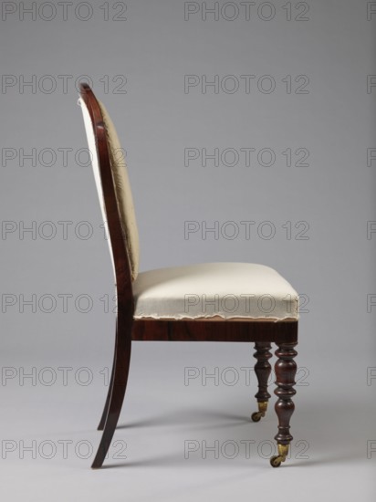 Chair with a padded balloon backrest and a trapezoidal padded seat removable cushion. Front legs are spool-turned, on brass rollers with clamps; rear legs are angular, tapered, slightly arched backwards, continuing into the backrest frames. Low frame with rounded front corners. Upholstery fabric not original, damask with a large floral pattern in purple and cream, finished with upholstery tape., Ca. 1840, Warsaw. Seating furniture; height 97.5 cm, width 48 cm, depth 53.7 cm., A side view of a wooden chair with a curved back and upholstered seat, showing a dark brown polished frame and a light beige fabric covering on the backrest and seat with some frayed edges visible; the front legs are turned and darker wood with brass caster fittings, the rear legs curve outward, and the background is a smooth neutral gray.