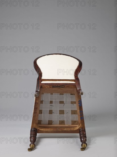 Chair with a padded balloon backrest and a trapezoidal padded seat removable cushion. Front legs are spool-turned, on brass rollers with clamps; rear legs are angular, tapered, slightly arched backwards, continuing into the backrest frames. Low frame with rounded front corners. Upholstery fabric not original, damask with a large floral pattern in purple and cream, finished with upholstery tape. ca. 1840, Warsaw. Seating furniture; height 97.5 cm, width 48 cm, depth 53.7 cm., The image shows the underside of a partially upholstered wooden chair or bench placed against a neutral gray background, with visible wooden frame in dark brown and lighter brown tones, a cream or off-white padded seat area, woven fabric webbing in light beige and off-white forming a lattice pattern, small touches of blue paint on the inner frame, brass or gold-colored caster wheels on turned dark brown wooden legs, and small exposed staples or stitching along the edge of the upholstery.