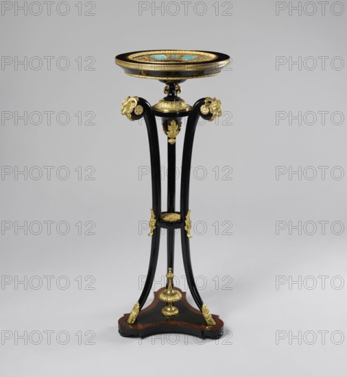 Round table, lacquered black, decorated with gilded cast brass fittings, with a ceramic painted bowl-shaped top supported by three slender channel-shaped supports connected in the middle by a round shelf, mounted on a triangular platform with concave sides. The platform has a profiled rim, with a central pinnacle a spinning top, on three low legs in the shape of strongly flattened spheres. The supports terminate in shoes at the bottom and in sheep's heads at the top. The tabletop is framed in a wooden, profiled frame on a spool-turned support, topped with a pine cone at the bottom and secured by a wooden hoop in the upper part of the supports. The porcelain tabletop plate is painted with overglaze paints, with the gold initial MG under a nine-baton crown in a trefoil field at the bottom, with brown tracery on a blue background border. Decoration includes goat's head motif, plant decoration, crowns, hooves animal legs, and monograms; styles noted: Empire and neo-Gothic. After 1860; like...