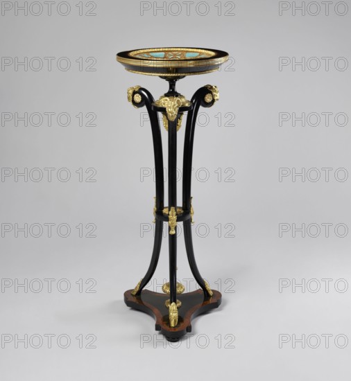 Business card table, revindication provenance, goat's head decoration, Empire and neo-Gothic styles, plant decoration, crowns, hooves animal legs, monograms. Round table lacquered black, decorated with gilded cast brass fittings, with a ceramic painted bowl-shaped top supported by three slender channel-shaped supports connected in the middle by a round shelf, mounted on a triangular platform with concave sides. The platform has a profiled rim, with a central pinnacle— a spinning top—on three low legs in the shape of strongly flattened spheres. The supports terminate in shoes at the bottom and in sheep's heads at the top. The tabletop is framed in a wooden profiled frame, on a spool-turned support, topped with a pine cone at the bottom, secured by a wooden hoop in the upper part of the supports. The porcelain tabletop plate is painted with overglaze paints, with the gold initial MG under a nine-baton crown in a trefoil field at the bottom, with brown tracery on a blue background border....