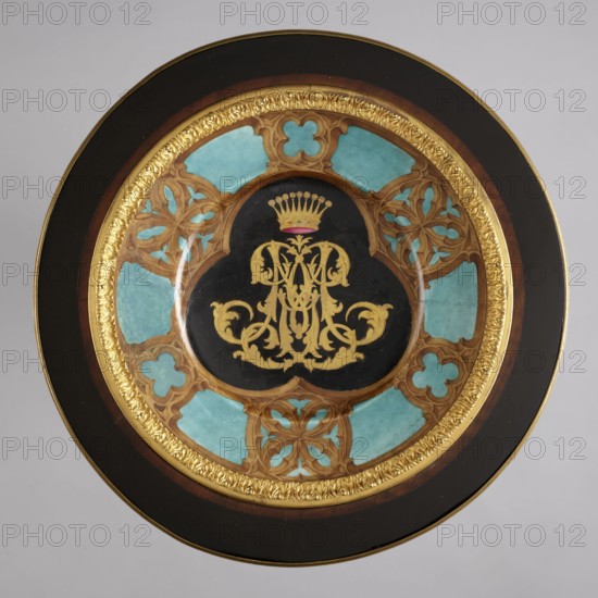 Round table, lacquered black, decorated with gilded cast brass fittings, with a ceramic painted bowl-shaped top supported by three slender channel-shaped supports connected in the middle by a round shelf, mounted on a triangular platform with concave sides. The platform has a profiled rim with a central pinnacle - a spinning top - on three low legs in the shape of strongly flattened spheres. The supports terminate in shoes at the bottom and in sheep's heads at the top. The tabletop is framed in a wooden profiled frame on a spool-turned support, topped with a pine cone at the bottom, secured by a wooden hoop in the upper part of the supports. The porcelain tabletop plate is painted with overglaze paints, with the gold initial MG under a nine-baton crown in a trefoil field at the bottom, with brown tracery on a blue background border. Decoration includes goat's head, plant motifs, crowns, hooves animal legs, and monograms; styles noted: Empire and neo-Gothic. After 1860; France origin? —...