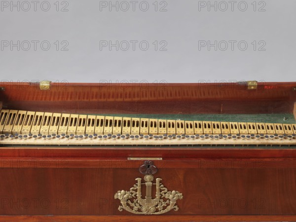 Harmonium by Jacob Deutschmann 1795–1853, 1822–1830, Vienna. Fisharmonia veneered with mahogany with a sliding mechanism and keyboard in an elongated rectangular case supported by two stringer legs. Signed inside the keyboard cover J. DEUTSCHMANN / IN WIEN brass sheet inlay. Legs and suspended pedal support lyre shaped like elongated vases. Forged openwork steel pedals. Keyboard table slides forward. Bone knobs on upper short sides lock the inner casing. Keyboard cover locks with a latch; braided hinges so-called piano. Biedermeier style; musical instrument. Dimensions: height 66 cm, width 100.6 cm, depth 27.9 cm., The image shows the inner section of a wooden musical instrument or case with a hinged top open to reveal a row of wooden key-like slats and circular felt or leather buttons, set against a strip of green felt; the wood has a reddish-brown finish with visible grain and patina, and there is an ornate metallic decorative emblem in a brass or gold tone mounted on the front panel...