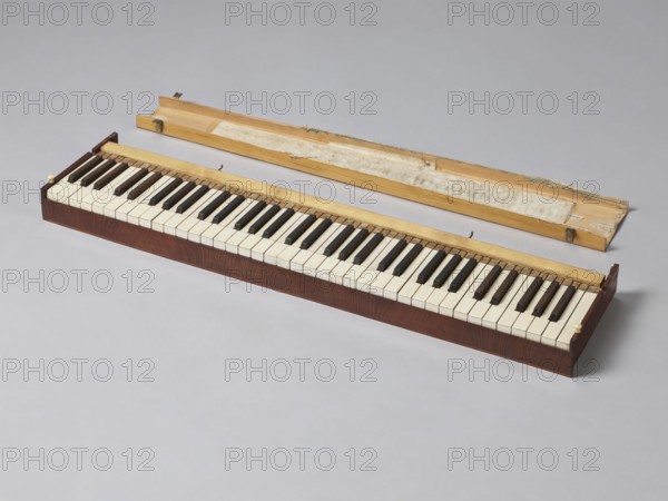 Harmonium, Jacob Deutschmann 1795–1853, 1822–1830. Fisharmonia veneered with mahogany, with a sliding mechanism and keyboard placed in an elongated rectangular case supported by two stringer legs; signed on the inside of the keyboard cover J. DEUTSCHMANN / IN WIEN brass sheet inlay. The legs and suspended pedal support lyre are in the shape of elongated vases. Forged openwork steel pedals. The keyboard table slides forward. Bone knobs on the upper part of the short sides lock the inner casing. Keyboard cover locks with a latch and braided hinges so-called piano. Biedermeier style. Vienna. Musical instrument; height 66 cm, width 100.6 cm, depth 27.9 cm., A long wooden keyboard module sits on a pale gray surface with its detached wooden lid placed behind it. The keyboard body is a dark brown wood with visible grain, topped by a lighter wood strip along the keys. The keys themselves are alternating light and dark colors, with the lighter keys appearing off-white and the darker keys appear...