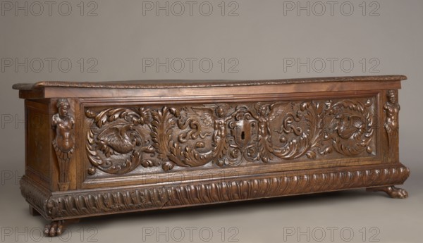 Chest, acanthus twig, fantastic animals, fantastic figures, snakes, coats of arms, herms decorative element, Renaissance style, animal paws, walnut. Cuboid carved chest supported by four low legs, the front ones in the shape of lion's paws, the rear ones angular. Front has an applied profiled frame enclosing a pseudo-panel carved with grotesque elements on a grained background: in the central section two winged genii merge at the bottom into an acanthus twig, holding a laurel wreath above a scrollwork cartouche framing the lock; between the volutes are twigs, snakes and griffins. Herms at the front corners. A prominent plinth molding is symmetrically decorated with gadroons. Straight side walls, originally with doorknobs. Interior on the left side of the cabinet. Hinges and lock striker of tinned, engraved steel. Ca. mid-16th century, Italy. Piece of furniture, storage unit. Height 54.5 cm, width 165.2 cm, depth 57 cm., A carved wooden chest displayed against a plain neutral background...