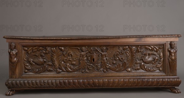 Chest, acanthus twig, fantastic animals, fantastic figures, snakes, coats of arms, herms decorative element, Renaissance style, animal paws, walnut. Chest, cuboid, carved, supported by four low legs, the front ones in the shape of lion's paws, the rear ones angular. On the front, an applied profiled frame enclosing a pseudo-panel carved with grotesque elements on a grained background: in the central section, two figures of winged genii, merging at the bottom into an acanthus twig, holding a laurel wreath above a scrollwork cartouche framing the lock; between the volutes, twigs, snakes and griffins. In the front corners, herms. A prominent plinth molding is symmetrically decorated with gadroons. Straight side walls, originally with doorknobs. Inside, on the left side of the cabinets. Hinges and lock striker made of tinned, engraved steel. Ca. mid-16th century, Italy, piece of furniture, storage unit; height 54.5 cm, width 165.2 cm, depth 57 cm., A carved wooden chest with a rich brown f...