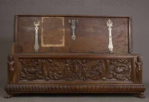Chest, repossession provenance noted, walnut. Cuboid carved storage chest, mid-16th century, Italy, Renaissance style. Supported by four low legs — front legs shaped as lion's paws, rear ones angular. Front has an applied profiled frame enclosing a pseudo-panel carved with grotesque elements on a grained background: central section shows two winged genii merging at the bottom into an acanthus twig, holding a laurel wreath above a scrollwork cartouche framing the lock; among the volutes are twigs, snakes, griffins, fantastic animals and figures, coats of arms, and herms decorative elements. Prominent plinth molding symmetrically decorated with gadroons. Straight side walls, originally with doorknobs. Hinges and lock striker of tinned, engraved steel. Function: piece of furniture/storage unit. Dimensions: height 54.5 cm, width 165.2 cm, depth 57 cm., A carved wooden chest with its lid open revealing the inside of the lid and the front panel, both showing a worn brown wood surface with me...