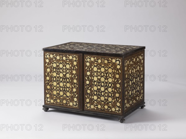 Cabinet, plant decoration, architectural columns, rosette ornament; early 20th century; India, and Europe ; piece of furniture storage; height 46.2 cm, width 63 cm, depth 43 cm., A rectangular wooden chest sits on small rounded feet against a pale background, showing ornate inlaid panels on its visible faces and top. The chest has a dark brown to nearly black ground with intricate floral and vine patterns in a contrasting pale gold or cream color covering the front doors, side, and top. The decorative panels feature repeating stylized flowers and curling leaves arranged within framed borders, and slim bands of lighter brown outline the panels. The feet and edges of the chest are the same dark color as the background, and the overall color palette includes dark brown or black, gold or cream, and some medium brown highlights.