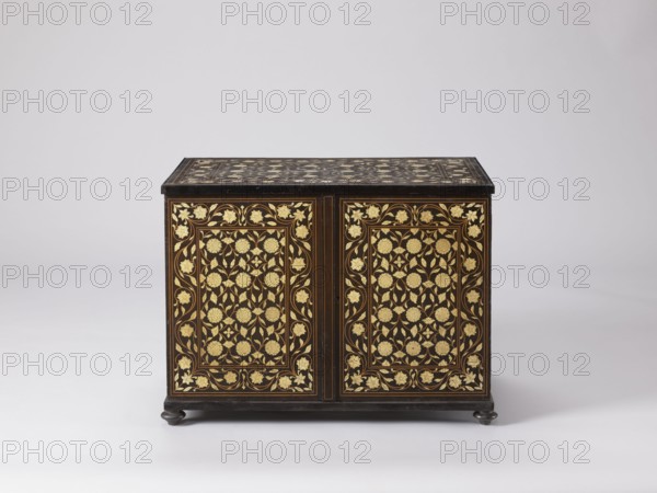 Cabinet, plant decoration, columns architectural, rosettes ornament, early 20th century, India possibly, Europe possibly, piece of furniture, storage piece; height 46.2 cm, width 63 cm, depth 43 cm., A rectangular wooden chest with a dark brown or black finish stands on small rounded feet; its front and top surfaces are decorated with an ornate floral pattern in a gold color set against the dark background, featuring stylized flowers and leaves arranged in repeating panels framed by thin borders, and the overall scene shows a few curved vines and floral motifs in gold contrasted with the dark wood, all presented on a pale neutral backdrop that is light gray to off-white.