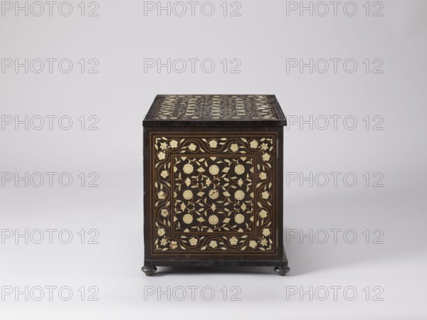 Cabinet, plant decoration, columns architectural, rosettes ornament, early 20th century, India, Europe, piece of furniture, storage piece; height 46.2 cm, width 63 cm, depth 43 cm, A dark wooden box with decorative inlay faces forward, showing an ornate floral pattern in light-colored material against the dark wood on its front and top surfaces; the box sits on small rounded feet and is photographed against a smooth, pale background in shades of white and light gray, with visible colors including dark brown or black of the wood and cream or pale gold of the inlaid floral motifs.