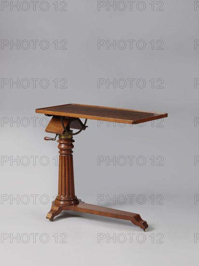 Bedside table, Biedermeier style; mahogany. Composed of an elongated platform outline similar to a triangle on three S-shaped legs with rollers; a fluted shaft mounted on it tapering upwards, and a rectangular top. The shaft is mounted on the short side of the platform, housing a prismatic column with a metal rack and a top swivel-mounted on it. The height of the top is adjusted using a crank working with the rack. The top can be tilted by a brass ring with a clamp that fixes its position. Turned wooden crank handle. 1815–1830, Europe. Furniture; table. Height 73.2 cm, width 81.2 cm, depth 41.2 cm., A wooden tilt-top table with a rectangular top in warm brown tones, supported by a turned, fluted central column and a single horizontal leg that ends in two carved feet with small brass castors; the table shows brass hardware where the top pivots and a small wooden handle on one side, all set against a plain light gray background with soft shadows.