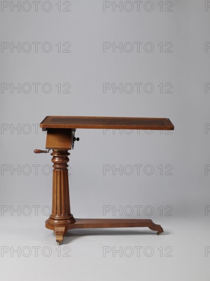 Bedside table, Biedermeier style, mahogany. Composed of an elongated platform outline similar to a triangle on three S-shaped legs with rollers; a fluted shaft mounted on it, tapering upwards, and a rectangular top. The shaft is mounted on the short side of the platform, housing a prismatic column with a metal rack and a top swivel-mounted on it. The height of the top is adjusted using a crank working with the rack. The top can be tilted by a brass ring with a clamp that fixes its position. Turned wooden crank handle. 1815–1830, Europe. Furniture; table. Height 73.2 cm, width 81.2 cm, depth 41.2 cm., A wooden adjustable table with a rectangular top in warm brown tones supported by a turned fluted central column and a horizontal stretcher base; the table shows a polished wood grain with darker and lighter brown variations, a small wooden crank handle on one side in a similar brown, brass or metal fittings at the crank and small caster wheels at the base with metallic tones, all set agai...