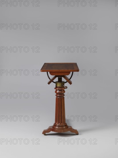Bedside table, Biedermeier style, mahogany. Composed of an elongated platform outline similar to a triangle on three S-shaped legs with rollers; a fluted shaft mounted on it, tapering upwards, and a rectangular top. The shaft is mounted on the short side of the platform, housing a prismatic column with a metal rack and a top swivel-mounted on it. The height of the top is adjusted using a crank working with the rack. The top can be tilted by a brass ring with a clamp that fixes its position. Turned wooden crank handle. 1815–1830, Europe. Furniture; table. Height 73.2 cm, width 81.2 cm, depth 41.2 cm., A wooden stand with a square top supported by a turned, fluted central column and a tripod base, shown against a plain light gray background; the stand displays warm brown and reddish-brown wood tones with brass-colored metal fittings and small dark caster wheels, and a curved dark metal support is visible under the top.