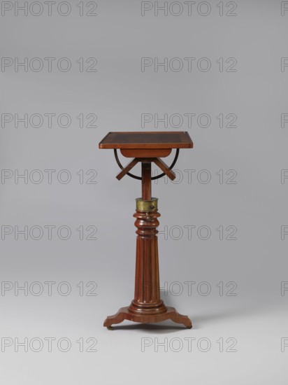 Bedside table, Biedermeier style, mahogany. Composed of an elongated platform outline similar to a triangle on three S-shaped legs with rollers; a fluted shaft mounted on it, tapering upwards, and a rectangular top. The shaft is mounted on the short side of the platform, housing a prismatic column with a metal rack and a top swivel-mounted on it. The height of the top is adjusted using a crank working with the rack. The top can be tilted by a brass ring with a clamp that fixes its position. Turned wooden crank handle. 1815–1830, Europe. Furniture; table. Height 73.2 cm, width 81.2 cm, depth 41.2 cm., A wooden pedestal table with a square top supported by a turned column and tripod base sits against a neutral grey background; the table shows a rich brown wood tone with darker brown accents on the top surface, a brass collar near the top of the column, and small brass casters at the feet, and there are curved and fluted details on the column and base, with a visible semicircular metal su...
