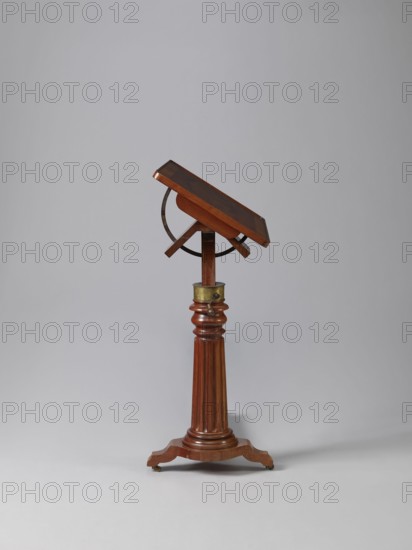 Bedside table, Biedermeier style, mahogany. Composed of an elongated platform outline similar to a triangle on three S-shaped legs with rollers; a fluted shaft mounted on it, tapering upwards, and a rectangular top. The shaft is mounted on the short side of the platform, housing a prismatic column with a metal rack and a top swivel-mounted on it. The height of the top is adjusted using a crank working with the rack. The top can be tilted by a brass ring with a clamp that fixes its position. Turned wooden crank handle. Date: 1815–1830. Place of origin: Europe. Furniture, table. Dimensions: height 73.2 cm; width 81.2 cm; depth 41.2 cm., A wooden adjustable lectern or stand with a rectangular slanted top in a polished dark brown wood held by a semicircular metal bracket, mounted on a turned fluted column of medium brown wood with a brass collar near the top and a carved tripod base on small casters, set against a plain light gray background; visible colors include dark brown, medium brown...