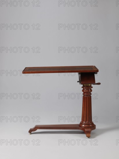 Bedside table, Biedermeier style, mahogany, composed of an elongated platform outline similar to a triangle on three S-shaped legs with rollers, a fluted shaft mounted on it tapering upwards, and a rectangular top. The shaft is mounted on the short side of the platform, housing a prismatic column with a metal rack and a top swivel-mounted on it. The height of the top is adjusted using a crank working with the rack. The top can be tilted by a brass ring with a clamp that fixes its position. Turned wooden crank handle. 1815–1830, Europe. Furniture; table. Height 73.2 cm; width 81.2 cm; depth 41.2 cm., A wooden side table with a rectangular top in rich brown wood finish supported by a turned fluted central column and a horizontal base; the table shows a few metal fittings near the top and small brass casters at the base, and the background is a smooth light gray.