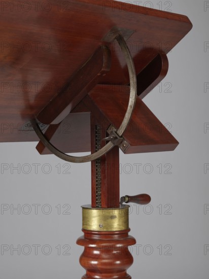 Bedside table, Biedermeier style, mahogany; composed of an elongated platform outline similar to a triangle on three S-shaped legs with rollers, a fluted shaft mounted on it tapering upward, and a rectangular top. Shaft mounted on the short side of the platform houses a prismatic column with a metal rack and a top swivel-mounted on it. Height of the top adjusted using a crank working with the rack; top can be tilted by a brass ring with a clamp that fixes its position. Turned wooden crank handle. Date: 1815–1830, Europe. Function: furniture table. Dimensions: height 73.2 cm, width 81.2 cm, depth 41.2 cm., The image shows a close-up of the underside of a wooden table or stand with a rich reddish-brown finish, featuring a carved turned wooden column in reddish-brown and a rectangular wooden support under the tabletop in similar reddish-brown tones; a curved metal band in a dull silvery-gray wraps beneath the tabletop and connects to a metal bracket attached to the wooden support, and a v...
