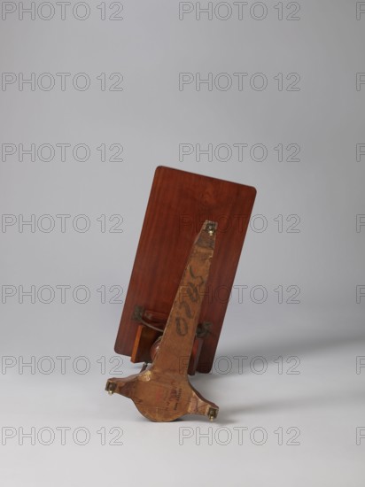 Bedside table, Biedermeier style, mahogany. Composed of an elongated platform outline similar to a triangle on three S-shaped legs with rollers; a fluted shaft mounted on it, tapering upwards, and a rectangular top. The shaft is mounted on the short side of the platform, housing a prismatic column with a metal rack and a top swivel-mounted on it. The height of the top is adjusted using a crank working with the rack. The top can be tilted by a brass ring with a clamp that fixes its position. Turned wooden crank handle. 1815–1830, Europe. Furniture; table. Height 73.2 cm, width 81.2 cm, depth 41.2 cm., A wooden object with a reddish-brown rectangular panel leaning at an angle is shown against a light gray background; attached to the panel is a worn wooden support arm with a darker brown patina and visible wood grain, ending in a rounded base with a few metal fittings and small brackets, and there are areas of lighter worn wood and darker scuff marks on the support.