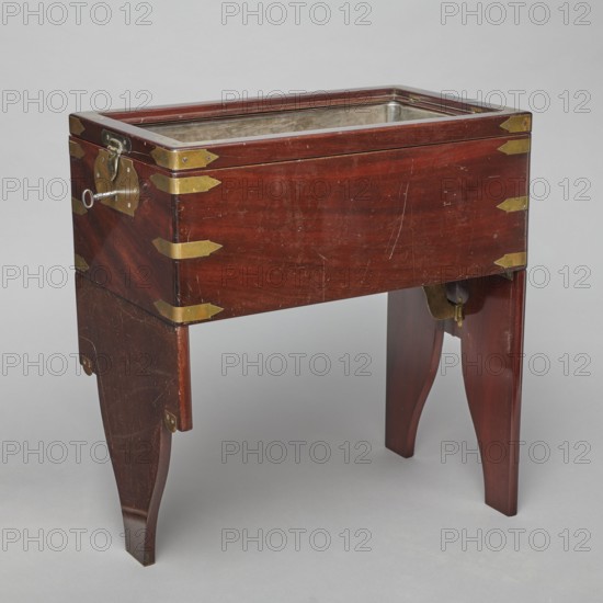 Bidet, unknown origin, hygiene; cuboid mahogany bidet containing a plated container of the same shape, closed with a removable lid; with folding supports. Hinged supports placed on the shorter sides with an S-shaped outline, forming a rectangular plate when folded the wider support on the inside is cut in the form of a concave-convex arch, filled with a narrower one of the same outline. Frame cover with a single removable panel. Brass fittings: strip fittings in the corners of the body three each and the lid single; sapphire hinges; a latch locking the supports; folding, rotating locks for both supports; and a pair of flat locks for the rear support. Removable container with extendable handles made of thick wire on long sides. 18th/19th century, England; furniture — toilet equipment; height 52.2 cm, width 47.7 cm, depth 28.5 cm., A wooden rectangular box with a reddish-brown finish stands on a pair of matching reddish-brown legs, showing a few scratches and worn areas on its surface; t...