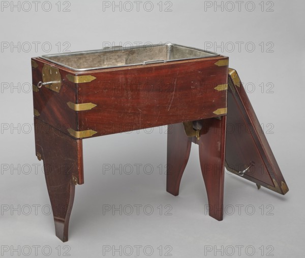 Bidet, unknown origin, hygiene; cuboid mahogany bidet containing a plated container of the same shape, closed with a removable lid; with folding supports. Hinged supports placed on the shorter sides with an S-shaped outline, forming a rectangular plate when folded the wider support on the inside is cut in the form of a concave-convex arch, filled with a narrower one of the same outline. Frame cover with a single removable panel. Brass fittings: strip fittings in the corners of the body three each and the lid single, sapphire hinges; a latch locking the supports, folding rotating locks for both supports, and a pair of flat locks for the rear support. Removable container with extendable handles made of thick wire. 18th/19th century, England; furniture, toilet equipment. Dimensions: height 52.2 cm, width 47.7 cm, depth 28.5 cm., A wooden rectangular washstand with a deep basin inset in the top sits on four tapered legs, with one triangular wooden panel detached and leaning against the rig...