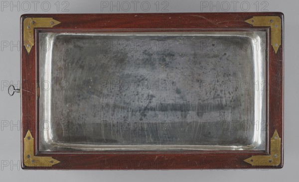 Bidet, unknown origin, hygiene; cuboid mahogany bidet containing a plated container of the same shape, closed with a removable lid; with folding supports. Hinged supports placed on the shorter sides with an S-shaped outline, forming a rectangular plate when folded the wider support on the inside is cut in the form of a concave-convex arch, filled with a narrower one of the same outline. Frame cover with a single removable panel. Brass fittings: strip fittings in the corners of the body three each and the lid single, sapphire hinges; a latch locking the supports, folding, rotating locks for both supports, and a pair of flat locks for the rear support. Removable container with extendable handles made of thick wire. 18th/19th century, England; furniture, toilet equipment. Height 52.2 cm, width 47.7 cm, depth 28.5 cm., A rectangular object with a polished dark brown wooden frame featuring a few brass corner brackets and a metal key protruding from one side encloses a reflective, slightly w...