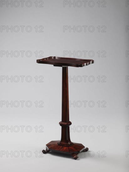Pedestal table with a rectangular top with a wavy-cut edge and a low wooden gallery, supported by an angular shaft widening toward the bottom, mounted on an octagonal platform on four volute legs. The shaft has lacing in the lower part; the platform is faceted., 1820–1835, England. Furniture table. Height 75.8 cm; width 39.5 cm; depth 27.5 cm., A wooden pedestal table with a dark brown finish stands against a light gray background; it has a rectangular top with a raised, scalloped edge and a glossy surface, a tapered central column with a rounded collar near the base, and an octagonal platform base resting on a group of small rounded feet.