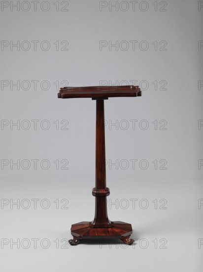Pedestal table with a rectangular top with a wavy-cut edge and a low wooden gallery, supported by an angular shaft widening toward the bottom, mounted on an octagonal platform on four volute legs. The shaft has a lacing in the lower part; the platform is faceted. Circa 1820–1835, England. Furniture table. Height 75.8 cm, width 39.5 cm, depth 27.5 cm., A tall, narrow wooden stand with a rectangular top that has raised, carved edges sits against a plain light gray background; the stand has a slender central column that tapers slightly and is finished in a rich reddish-brown wood grain, a rounded collar about midway, and an octagonal base of the same wood with carved, dark brown claw-like feet.