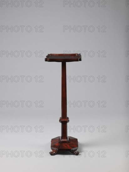Pedestal table with a rectangular top with a wavy-cut edge and a low wooden gallery, supported by an angular shaft widening towards the bottom, mounted on an octagonal platform on four volute legs. The shaft has a lacing in the lower part; the platform is faceted. 1820–1835, England. Furniture table. Height 75.8 cm; width 39.5 cm; depth 27.5 cm., A tall, narrow wooden stand with a dark reddish-brown finish, featuring a rectangular top with slightly upturned edges, a slender central column with a rounded midsection, and an octagonal base resting on a group of carved feet; the background is a smooth light gray.