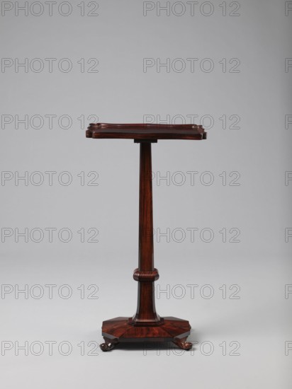 Pedestal table with a rectangular top with a wavy-cut edge and a low wooden gallery, supported by an angular shaft widening toward the bottom, mounted on an octagonal platform on four volute legs. The shaft has a lacing in the lower part; the platform is faceted. Makers unknown. 1820–1835, England. Furniture; table. Height 75.8 cm, width 39.5 cm, depth 27.5 cm., A wooden pedestal-style stand with a rectangular top that has slightly raised and carved edges, supported by a single tapered central column with a rounded collar detail midway, resting on an octagonal base with carved feet. The colors visible are deep reddish-brown and dark brown wood tones against a light gray background.