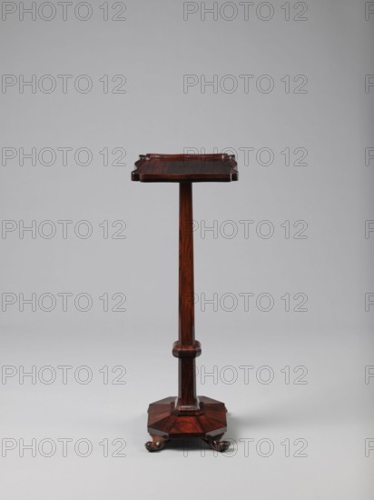 Pedestal table with a rectangular top with a wavy-cut edge and a low wooden gallery, supported by an angular shaft widening towards the bottom, mounted on an octagonal platform on four volute legs. The shaft has lacing in the lower part; the platform is faceted., 1820–1835, England. Furniture; table. Height 75.8 cm, width 39.5 cm, depth 27.5 cm., A tall wooden stand with a dark reddish-brown finish, featuring a square top with scalloped edges, a slender central column, a rounded mid-rail detail, and an octagonal base resting on carved paw feet; the background is a smooth light gray.