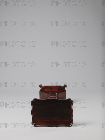 Pedestal table with a rectangular top with a wavy-cut edge and a low wooden gallery, supported by an angular shaft widening toward the bottom, mounted on an octagonal platform on four volute legs. The shaft has lacing in the lower part; the platform is faceted., 1820–1835, England. Furniture; table. Height 75.8 cm, width 39.5 cm, depth 27.5 cm., A polished dark reddish-brown wooden object with a wavy rectangular front face sits on a pale gray surface against a light gray background, showing wood grain patterns and a glossy finish; behind it a matching wooden section with angular facets and small carved rounded protrusions is partially visible in similar dark reddish-brown tones.