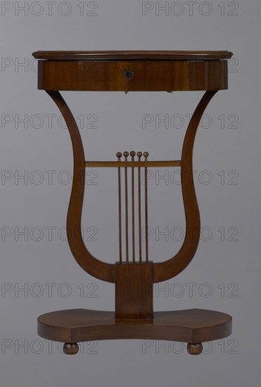 Oval display table with lyre-shaped base mounted on a platform with a concave-convex outline, supported by four spherical legs. Veneered with mahogany. Lyre strings terminate at the top with balls. Edge of the tabletop frame is profiled. Circa 1820–1840, Central Europe. Piece of furniture for displaying objects. Dimensions: height 76.7 cm; width 52.3 cm; depth 37.9 cm., A wooden lyre-shaped table with a rich brown finish set against a neutral gray background; the top is an oval wooden surface with a slightly raised edge and a darker brown hue, supported by curved side supports that form the lyre shape, connecting to a rectangular central post; a horizontal bar across the lyre holds a set of vertical thin wooden rods topped with small rounded finials, all in matching brown tones; the base is a rounded, contoured wooden platform on small rounded feet, also brown.