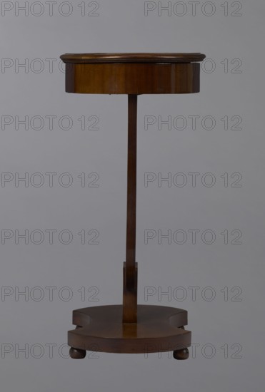 Oval display table with a lyre-shaped base mounted on a platform with a concave-convex outline, supported by four spherical legs. Entirely veneered in mahogany; the lyre strings terminate at the top with balls. The edge of the tabletop frame is profiled. Lyres musical-instrument motif. Piece of furniture for displaying objects., Dated 1820–1840, Central Europe. Dimensions: height 76.7 cm; width 52.3 cm; depth 37.9 cm., A slender wooden stand with a round top and a circular base connected by a straight central column, all in shades of brown against a plain gray background; the top has a slightly darker brown rim and a polished surface, the column is a medium brown, and the base sits on small rounded feet in darker brown.