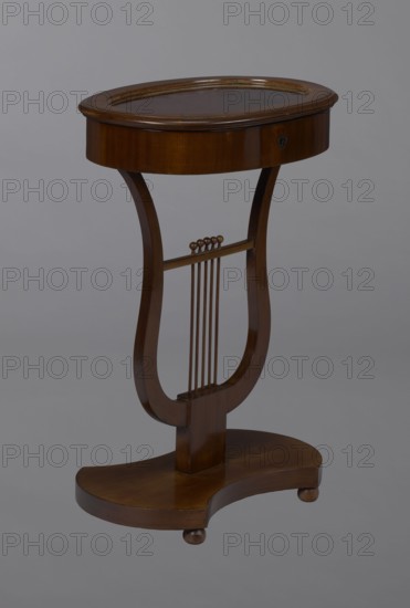 Oval display table with lyre-shaped base mounted on a platform with a concave-convex outline, supported by four spherical legs; veneered in mahogany. Lyre strings terminate at the top with balls. The edge of the tabletop frame is profiled. Display case table, purchase provenance. Lyres musical-instrument motif. 1820–1840, Central Europe. Piece of furniture for displaying objects. Height 76.7 cm; width 52.3 cm; depth 37.9 cm., A wooden side table with a rounded oval top and a shallow rim sits on a sculpted lyre-shaped support that connects to a curved base; the table has a polished brown finish with visible wood grain, a darker brown inset on the top surface, and small rounded feet beneath the base, all shown against a plain gray background.