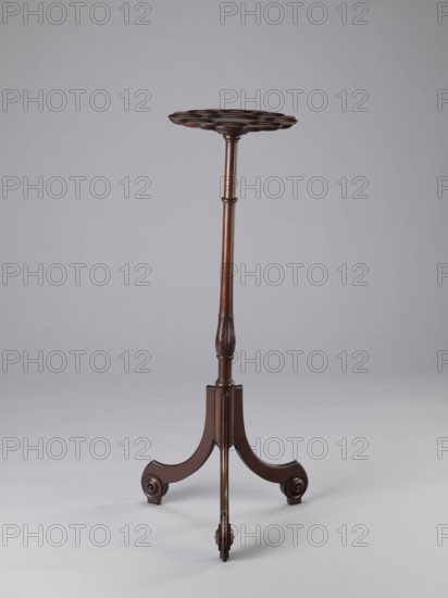 Side table, TABLE with a top in the form of a round saucer mounted on a turned stem forked into a tripod. The top has cut-out recesses for a jug and eight glasses. The central stem is in the form of a baluster with carved acanthus leaves. Legs made of channel-shaped slats, topped with volutes at the bottom, with turned knobs applied on the sides. 1820–1840, England. Furniture; table. Height 100.5 cm; width 39 cm; depth 39 cm., A tall, slender wooden stand with a dark brown finish, featuring a round scalloped top supported by a long turned central column that tapers toward an intricately carved section, ending in a tripod base with three curved legs each terminating in rounded scroll-like feet; the background and floor are shades of light gray.