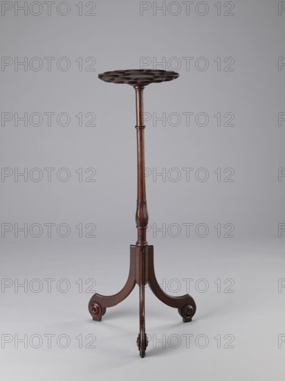 Side table, TABLE with a top in the form of a round saucer mounted on a turned stem forked into a tripod. The top has cut-out recesses for a jug and 8 glasses. The central stem is in the form of a baluster with carved acanthus leaves. Legs made of channel-shaped slats, topped with volutes at the bottom, with turned knobs applied on the sides. 1820–1840, England. Furniture, table. Height 100.5 cm; width 39 cm; depth 39 cm., A wooden stand with a round scalloped top is shown against a plain light gray background; the stand has a slender turned central column with carved details and a dark brown finish, ending in a tripod base with three curved legs, each leg terminating in a rounded foot with circular carved accents; visible colors include dark brown and lighter brown tones on the wood and light gray in the background.