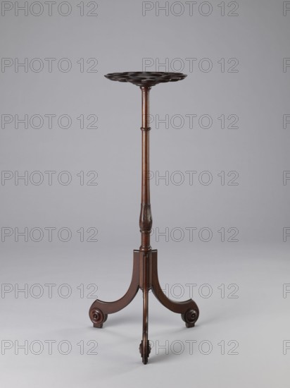 Side table, TABLE with a top in the form of a round saucer mounted on a turned stem forked into a tripod. The top has cut-out recesses for a jug and eight glasses. The central stem is in the form of a baluster with carved acanthus leaves. Legs made of channel-shaped slats, topped with volutes at the bottom, with turned knobs applied on the sides. 1820–1840, England. Furniture; table. Height 100.5 cm, width 39 cm, depth 39 cm., A tall, slender wooden stand with a dark brown finish features a round, scalloped top supported by a turned central column that narrows toward the middle and has carved detailing near the lower section; the base consists of a tripod of curved legs ending in scroll-like feet, all set against a pale gray background.