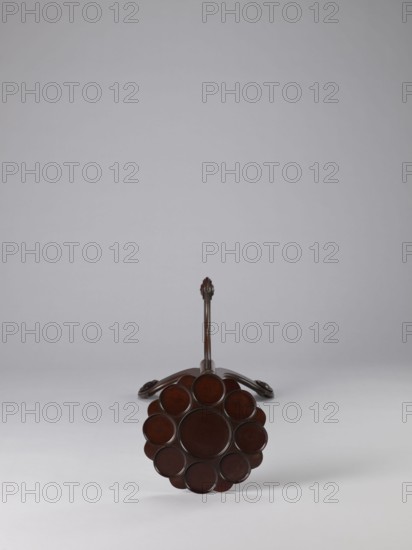 Side table, TABLE with a top in the form of a round saucer mounted on a turned stem forked into a tripod. The top has cut-out recesses for a jug and eight glasses. The central stem is in the form of a baluster with carved acanthus leaves. Legs made of channel-shaped slats, topped with volutes at the bottom, with turned knobs applied on the sides. 1820–1840, England. Furniture; table. Height 100.5 cm, width 39 cm, depth 39 cm., A dark brown wooden object viewed from the end showing a circular flower-like arrangement of round, slightly recessed discs forming a central circle surrounded by a ring of several smaller circles, with a long slender shaft extending back to a curved base that rests on a pale gray surface against a light gray background; visible colors include dark brown, pale gray, and light gray.